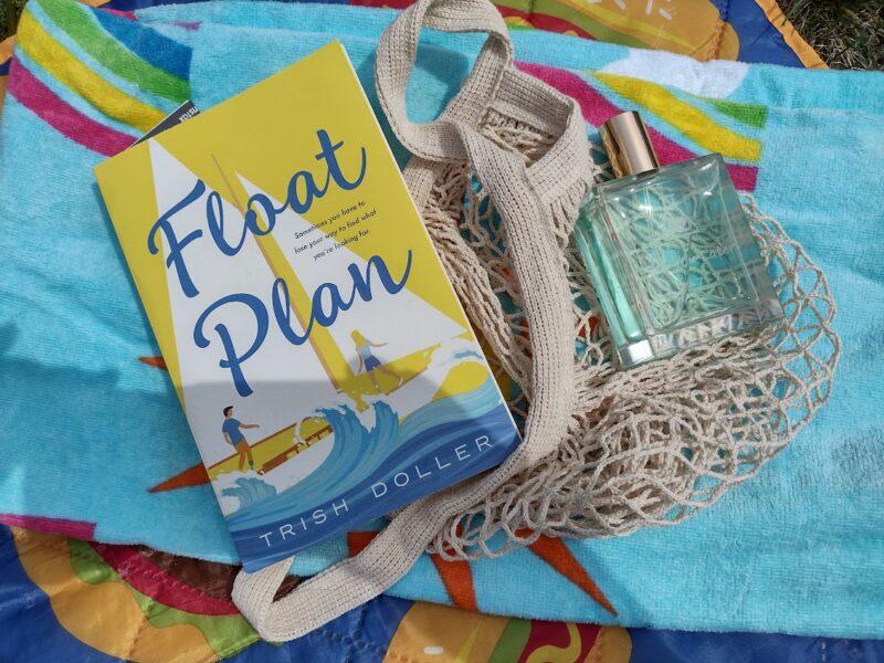 paperback copy of Float Plan on a beach towel with a bottle of aqua perfume and a net bag